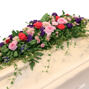 Blommor i olika färger, inklusive rosa och röda rosor samt lila blommor, arrangerade ovanpå en kista täckt med vit duk.